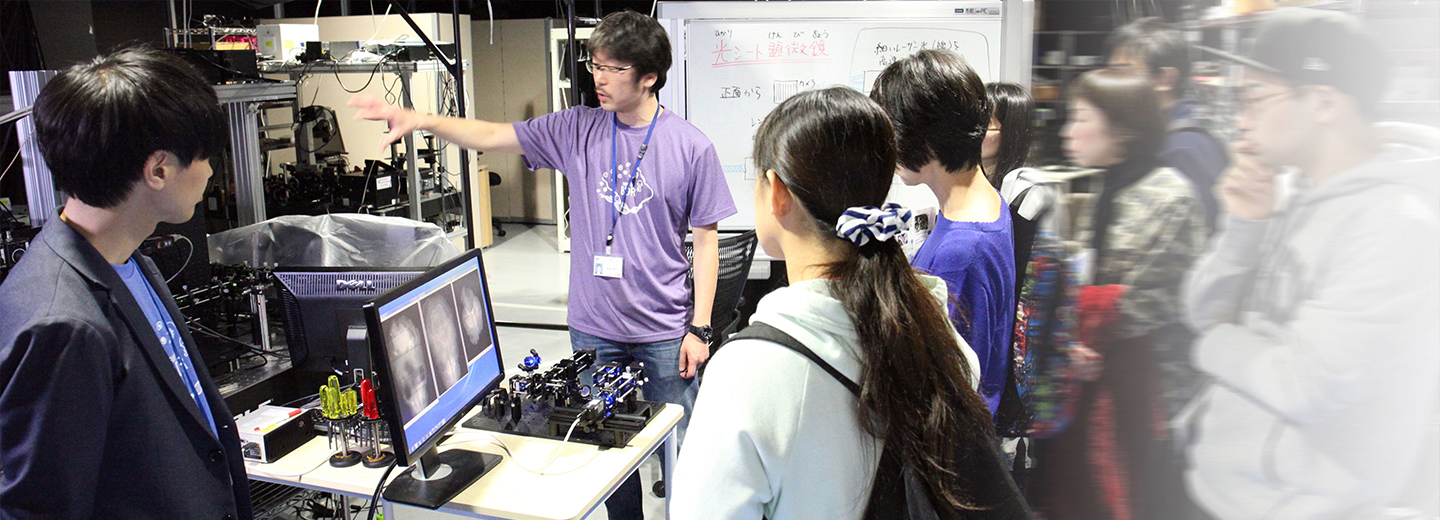 大阪一般公開で研究機器の説明をする研究者と来場者たちの写真